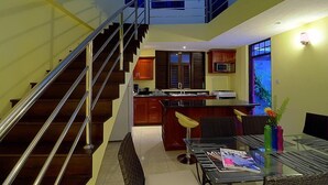 A modern interior with a staircase leading to an upper level, a kitchen with dark wooden cabinets, a dining area with a glass table, and a small island with a microwave.