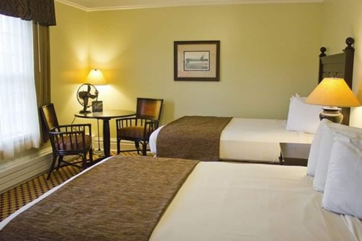 A hotel room with two beds, a small table with a lamp, a chair, and a framed picture on the wall.