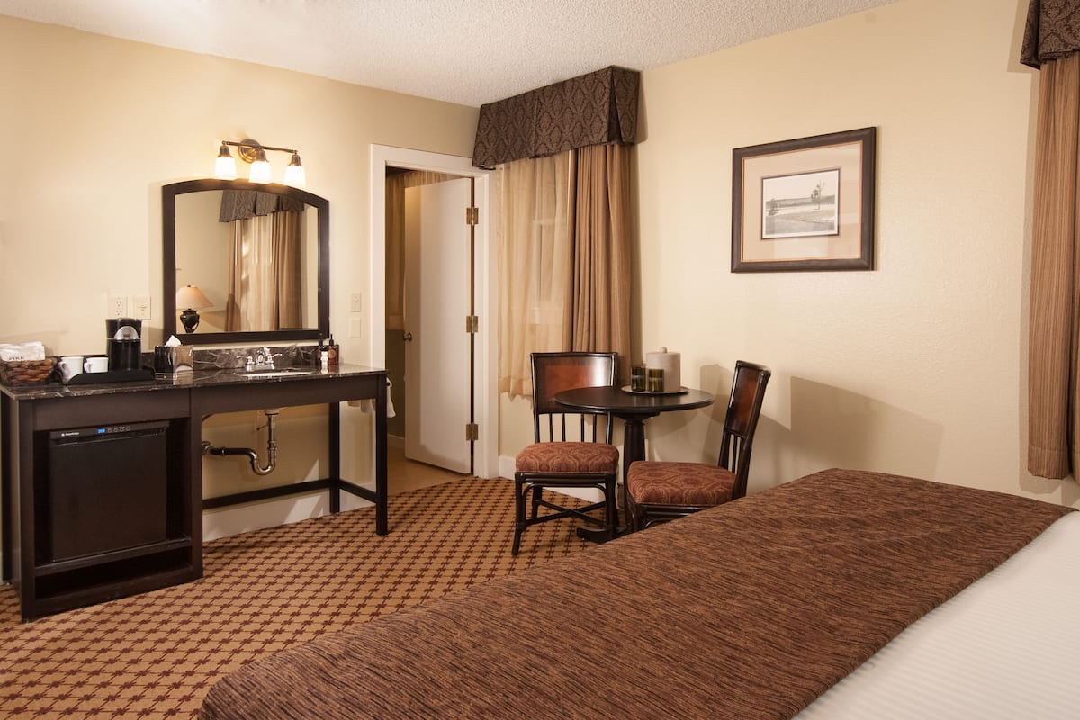 A hotel room with a bed, a desk with a mirror, a chair, a small table, and a framed picture on the wall.