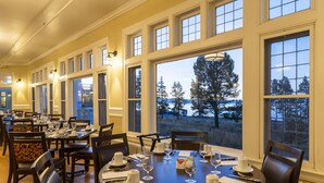 A dining area with multiple tables set for service, large windows offering a view of trees and a body of water, and a classic interior design with wooden furniture and white walls.