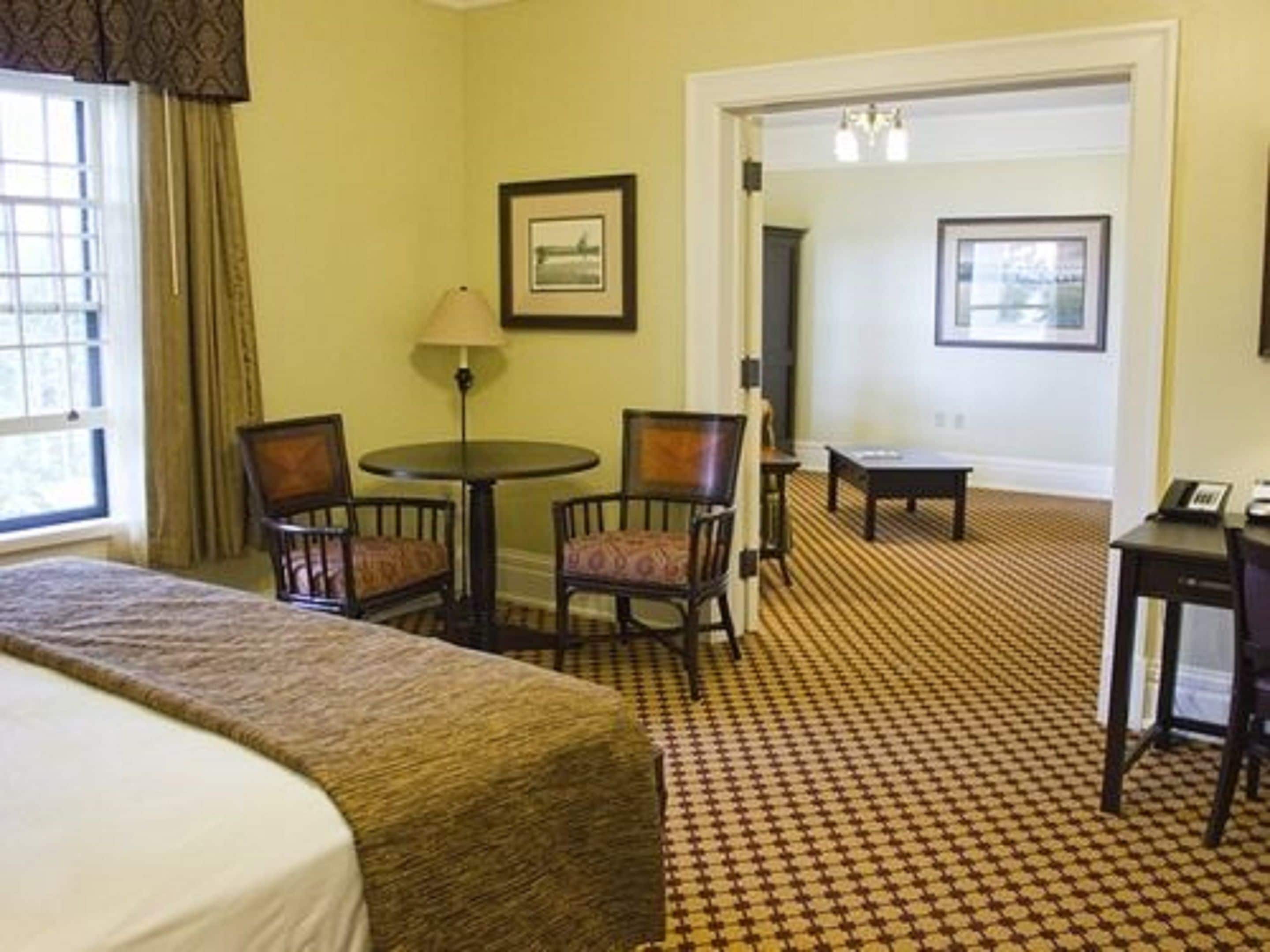A hotel room with a bed, two chairs, a small table, a lamp, and a framed picture on the wall.