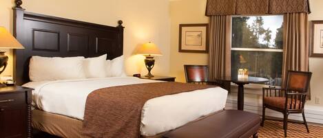 A hotel room with a large bed, two bedside lamps, a small table with a chair, and a window with curtains.