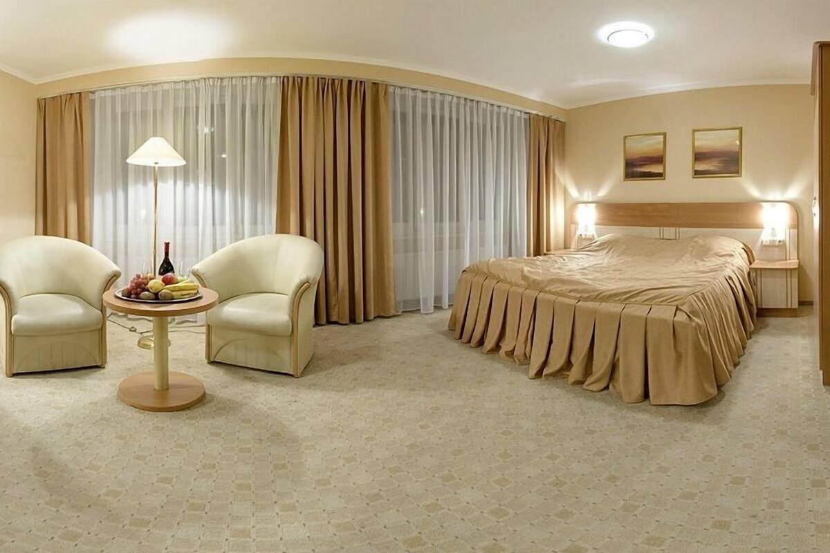 Ein Hotelzimmer mit einem Bett, zwei Sesseln, einem kleinen Tisch mit Früchten und einer Flasche sowie einem großen Fenster mit Vorhängen.