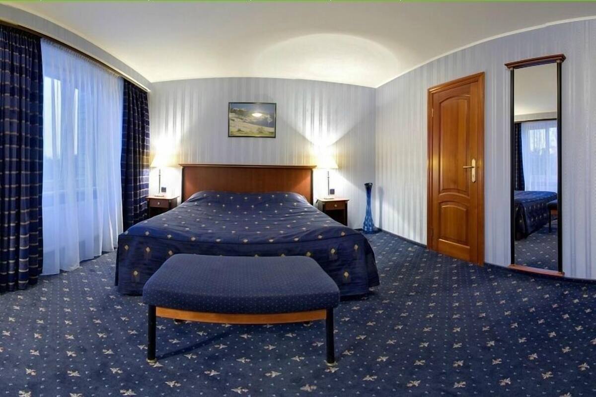 Ein Hotelzimmer mit einem großen Bett, einer Bank, einem Nachttisch, einer Lampe, einem Spiegel und einer Tür.