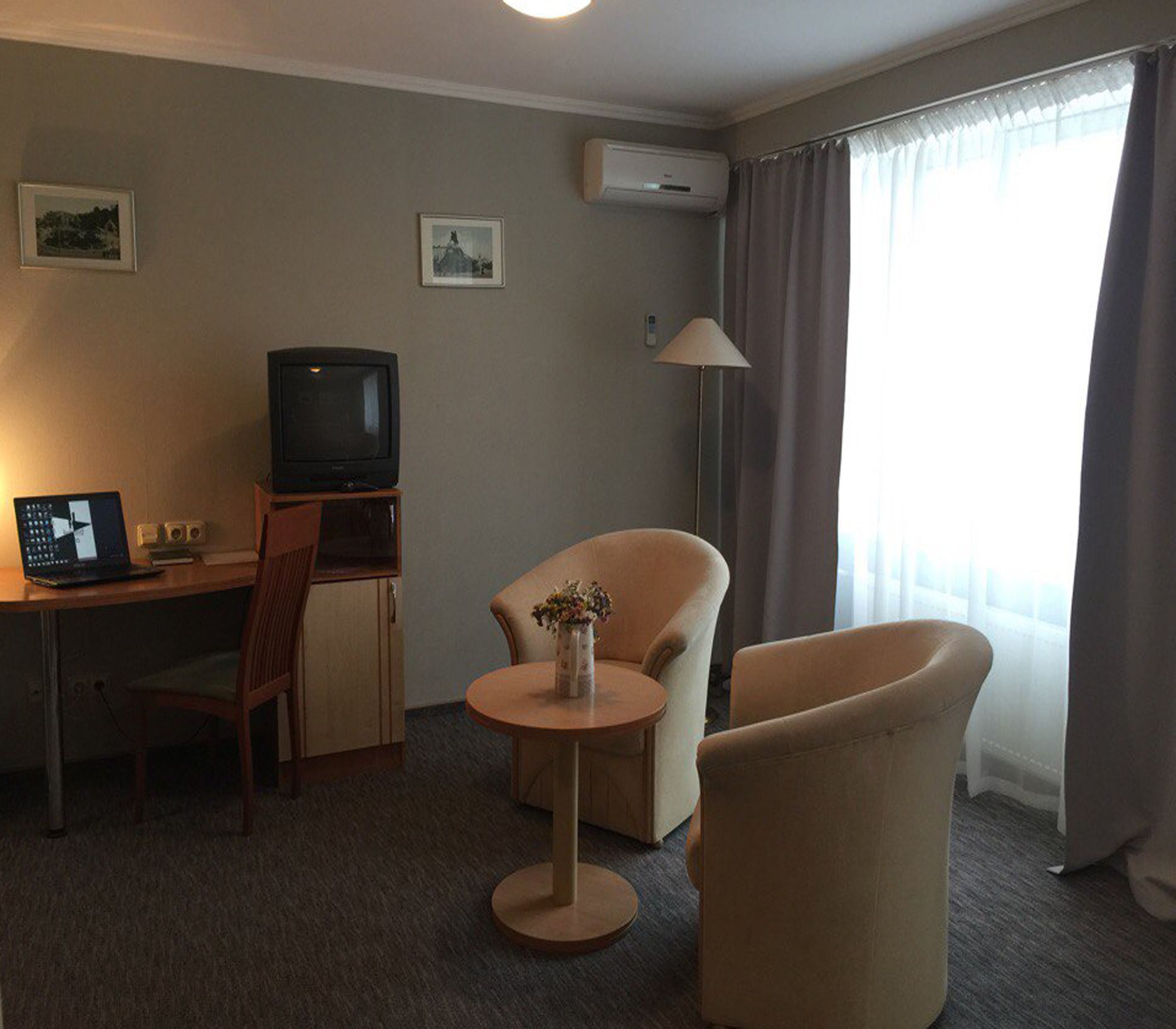 A hotel room with a desk, a television, a laptop, a chair, a round table with a vase, and a window with curtains.