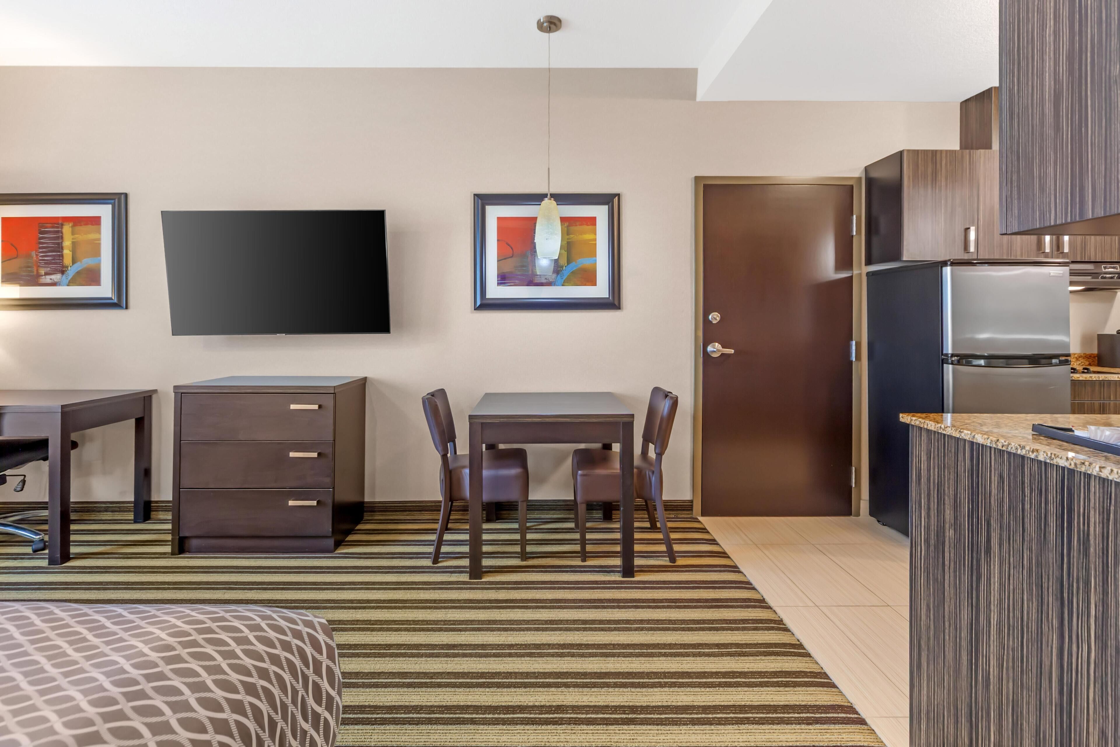 Quarto de hotel moderno com cama, escrivaninha com gavetas, TV, obras de arte emolduradas, mesa de jantar com cadeiras e cozinha equipada com frigorífico, além de uma porta.
