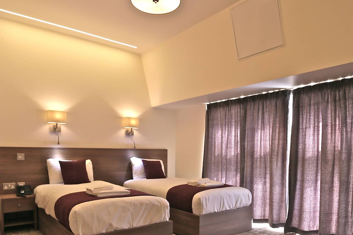 A hotel room with two beds, a nightstand, a lamp, and a large window with curtains.