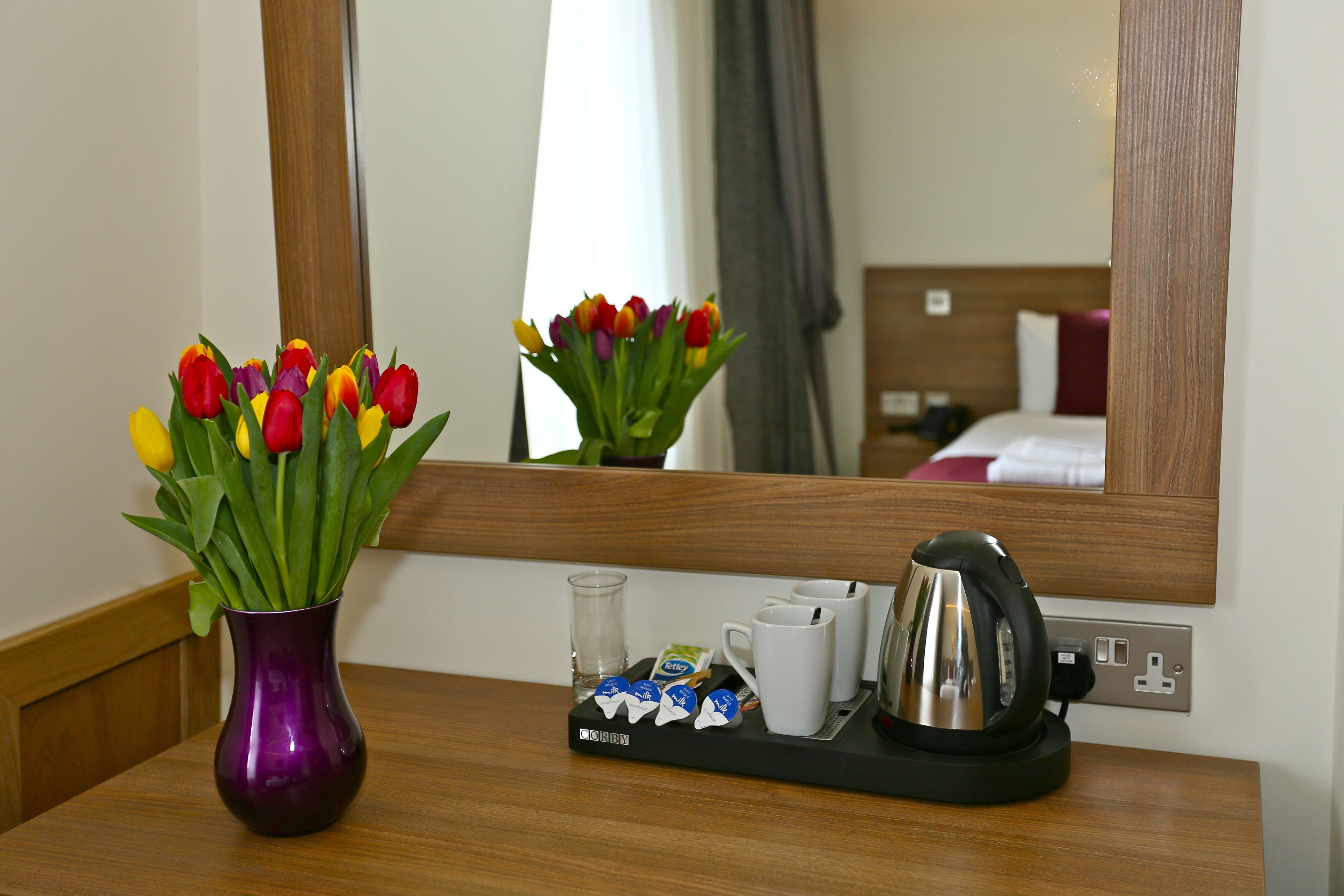 A hotel room with a wooden nightstand, a purple vase with tulips, a tea kettle, a tray with tea bags and a glass, and a mirror reflecting the room.