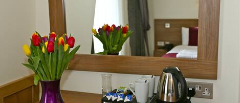A hotel room with a wooden nightstand, a purple vase with tulips, a tea kettle, a tray with tea bags and a glass, and a mirror reflecting the room.