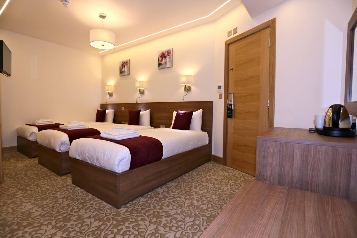 A hotel room with three beds, a wooden headboard, a desk with a coffee maker, and wall-mounted lamps.