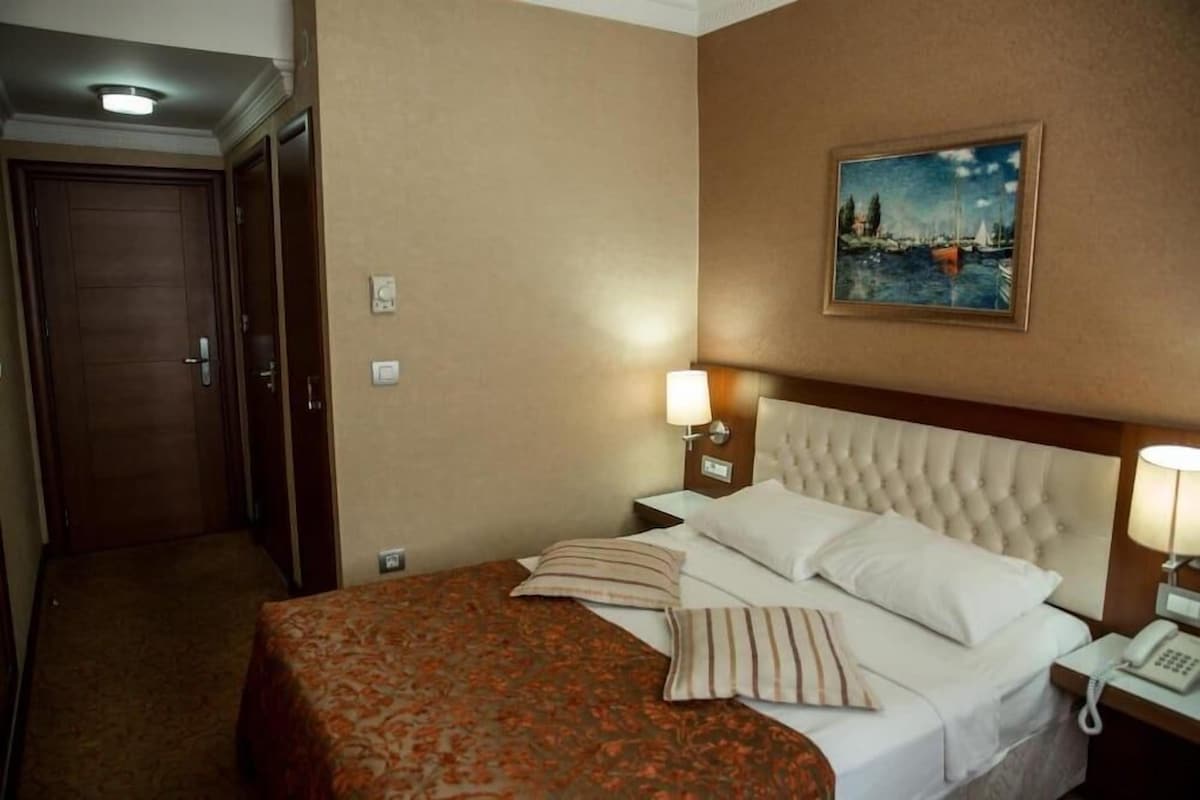 Una habitación de hotel con una cama, una mesita de noche, un teléfono, un cuadro en la pared y una puerta.