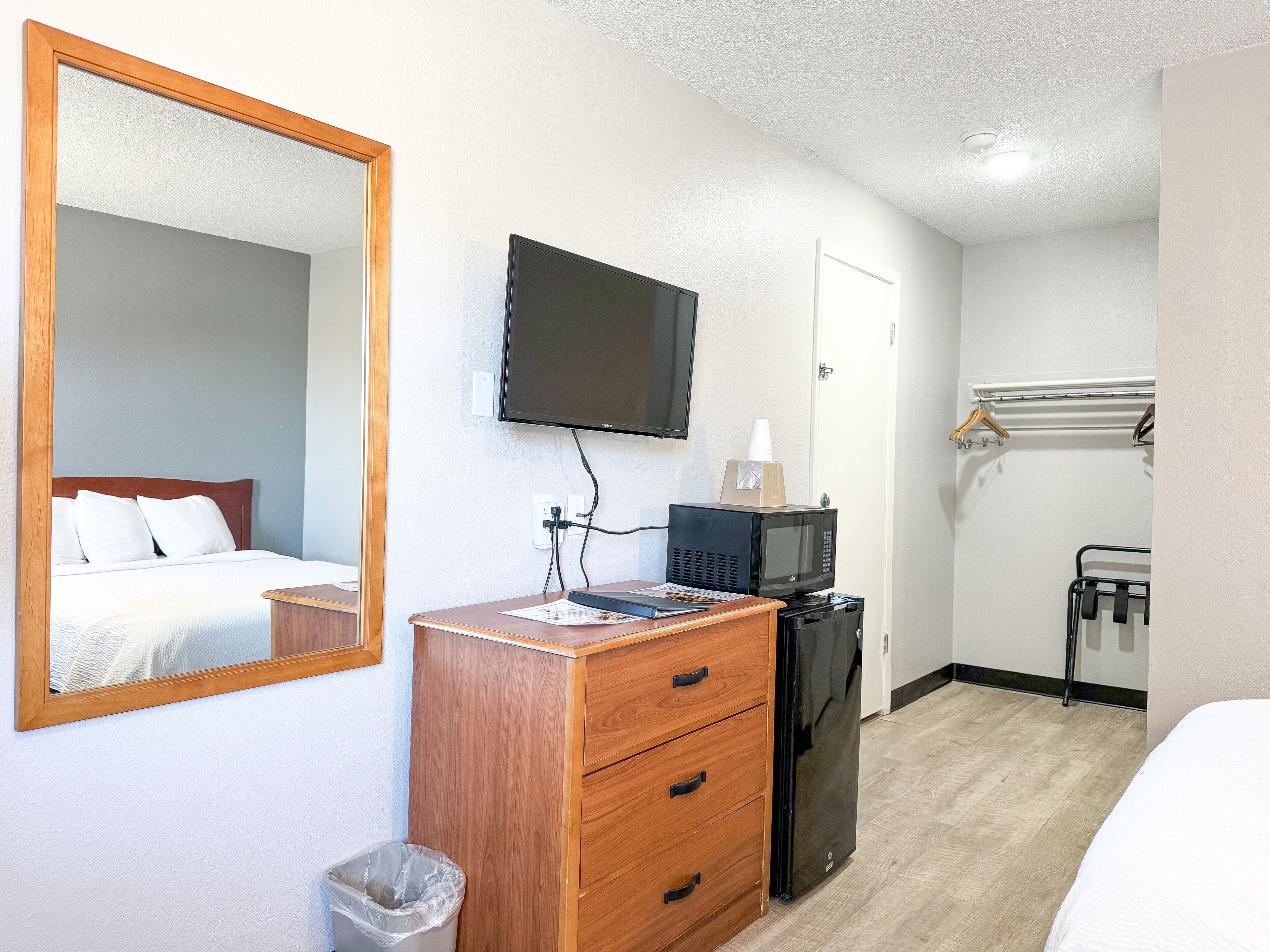Een hotelkamer met een houten dressoir, een spiegel, een flatscreen televisie, een magnetron, een kleine prullenbak en een bed met wit beddengoed.