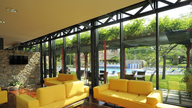 A modern living room with yellow sofas, a stone wall, and large windows overlooking a pool and garden. - Residence i Cormorani (Loano)