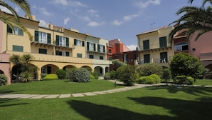 A courtyard with multiple buildings, green lawns, and palm trees. - Residence i Cormorani (Loano)