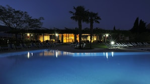 A nighttime view of a pool area with lounge chairs and a building with illuminated windows. - Residence i Cormorani (Loano)