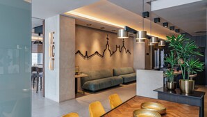 A modern interior with a dining area featuring a wooden table and yellow chairs, a grey sofa, and a wall with a geometric light fixture. - Vincci Mercat Hotel (Valencia)