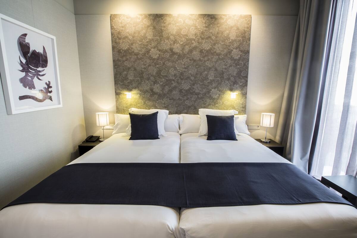 Uma cama de casal com lençóis brancos e almofadas decorativas escuras, um cabeceira estampada, abajures laterais e uma obra de arte emoldurada na parede.