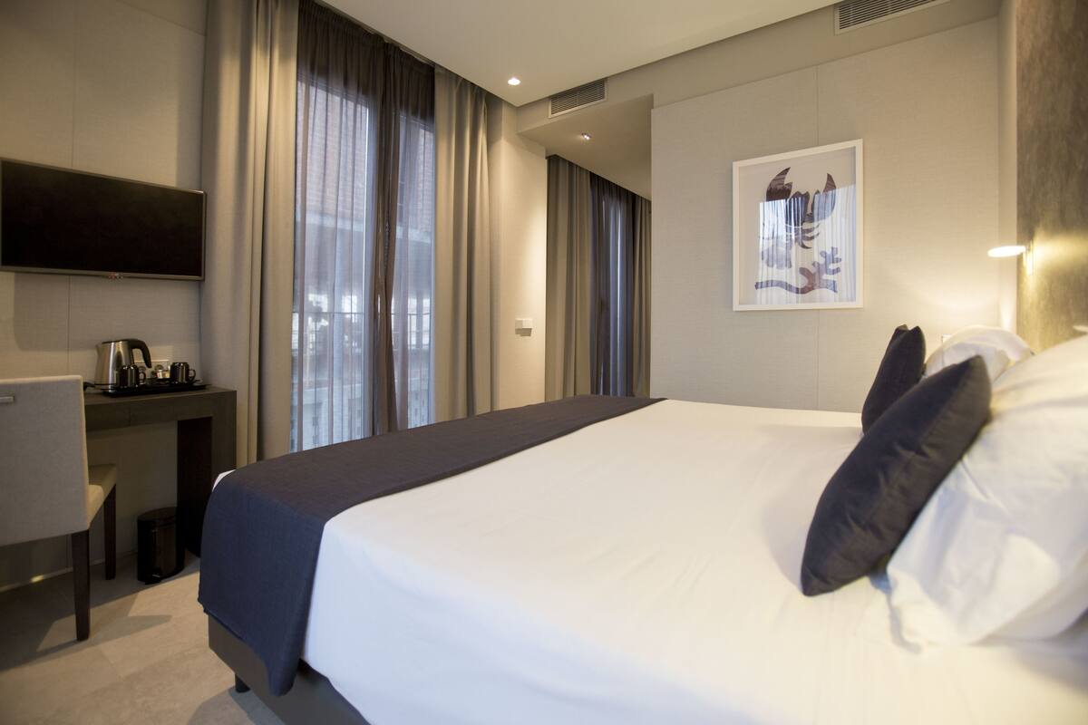 Quarto de hotel com uma cama grande, uma mesa com cafeteira, uma TV na parede e um quadro enquadrado na parede.