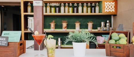 A bar area with a martini glass, a cocktail, and a menu sign. There are wooden crates with fruits and a potted plant.