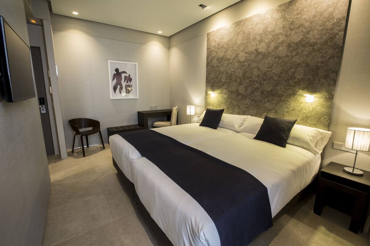 Quarto de hotel moderno com uma cama grande, mesas de cabeceira, escrivaninha e uma cadeira.