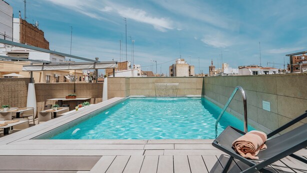 A rooftop pool with a view of the city skyline, a lounge chair with a towel, and outdoor seating. - Vincci Mercat Hotel (Valencia)