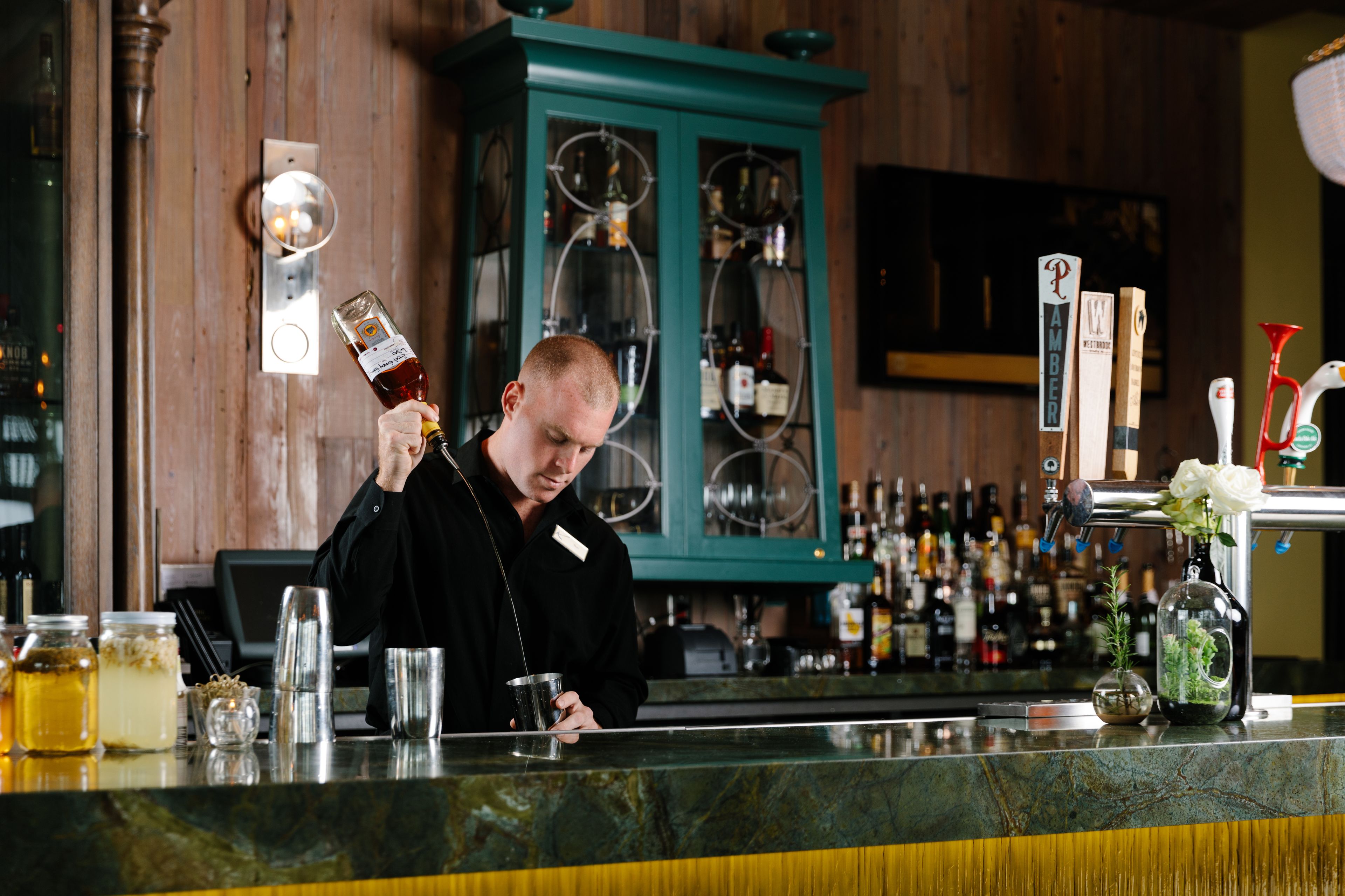 Un espace bar avec un barman tenant une bouteille, un comptoir avec diverses bouteilles et verres, et un meuble turquoise en arrière-plan.