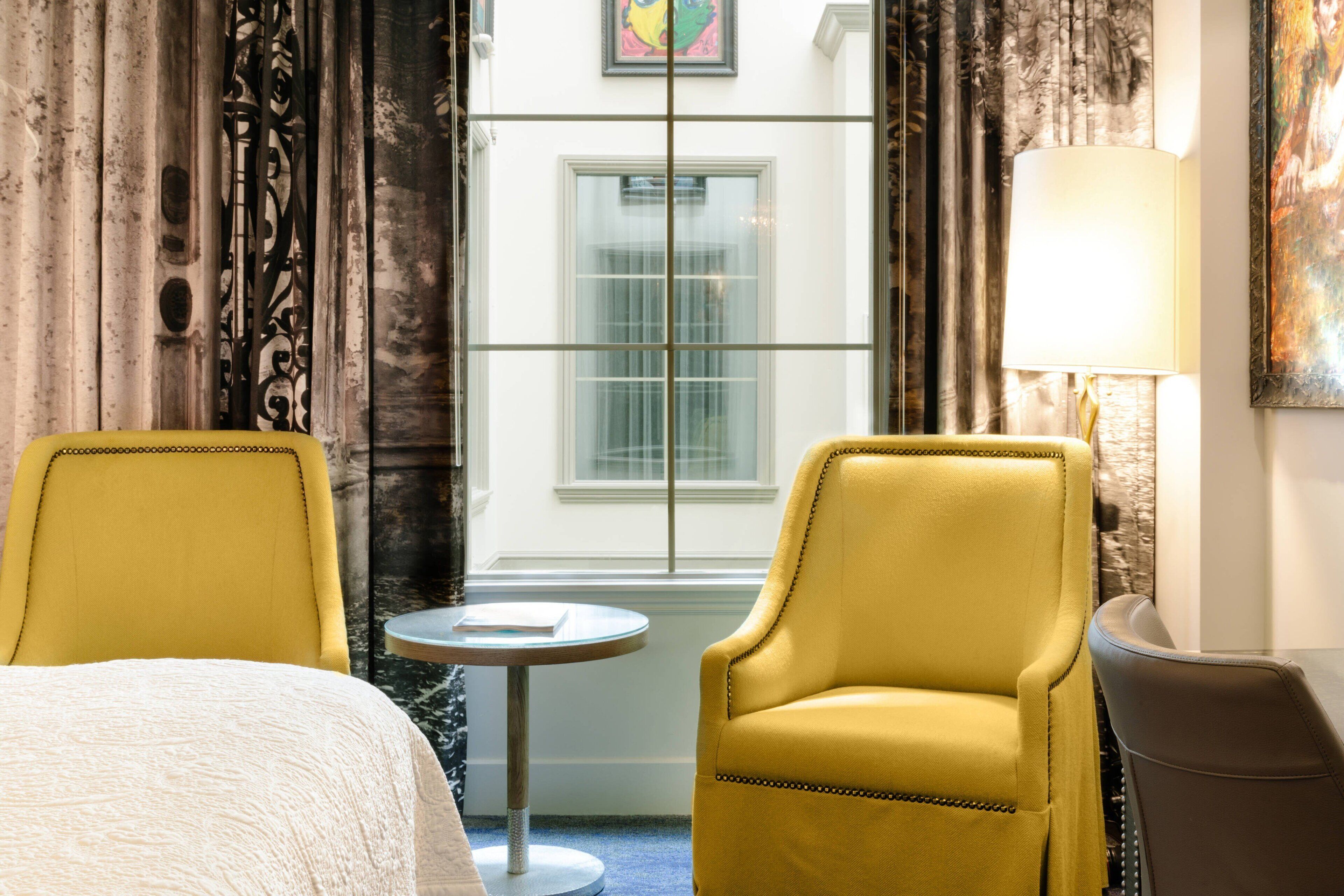 Une chambre d’hôtel avec un lit, deux fauteuils jaunes, une petite table ronde et une lampe.