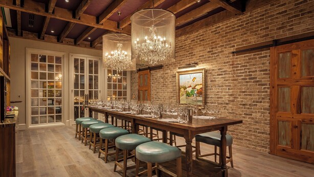 A long wooden bar with stools, a brick wall, and chandeliers.