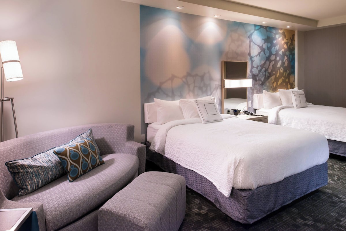 A hotel room with two beds, a sofa, a lamp, and a wall with abstract art.
