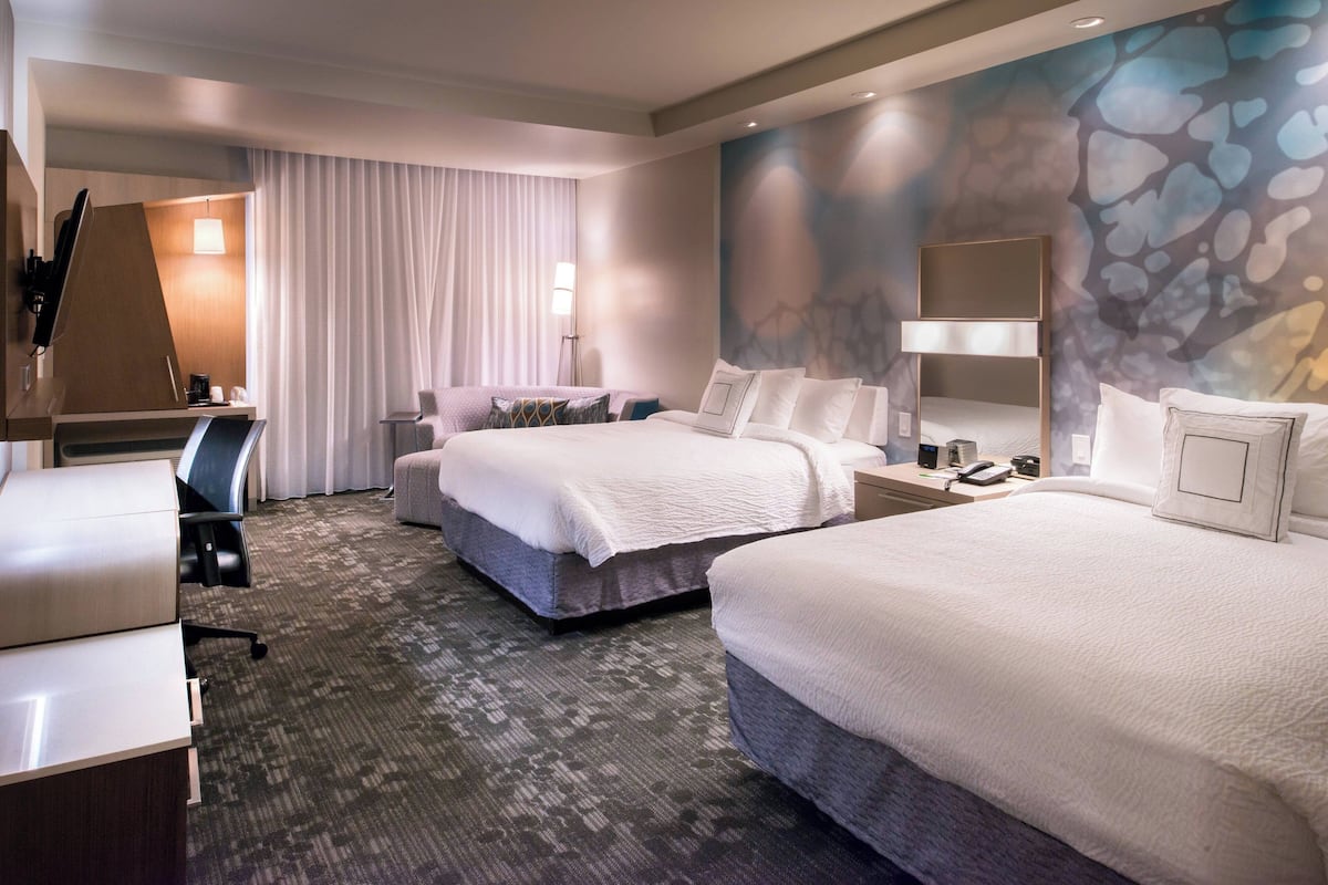 A hotel room with two beds, a desk, a chair, a lamp, and a large abstract wall mural.