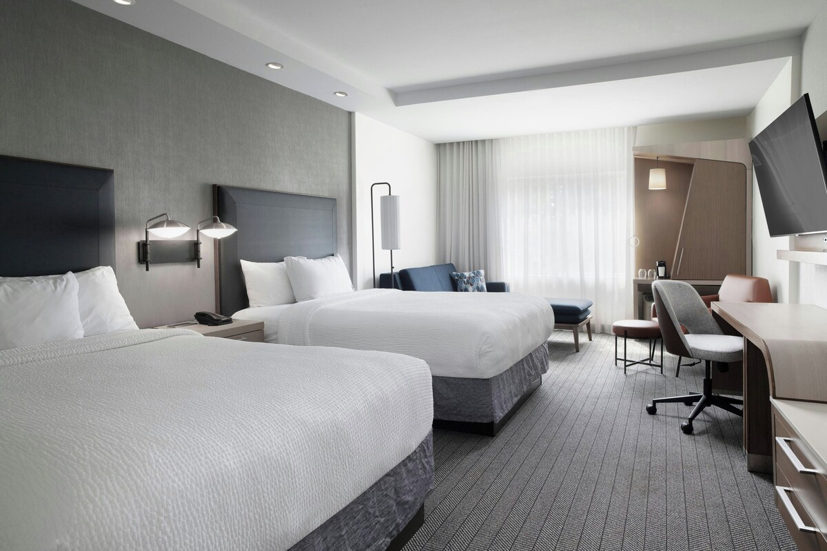 A hotel room with two beds, a desk, a chair, a TV, and a large window with curtains.