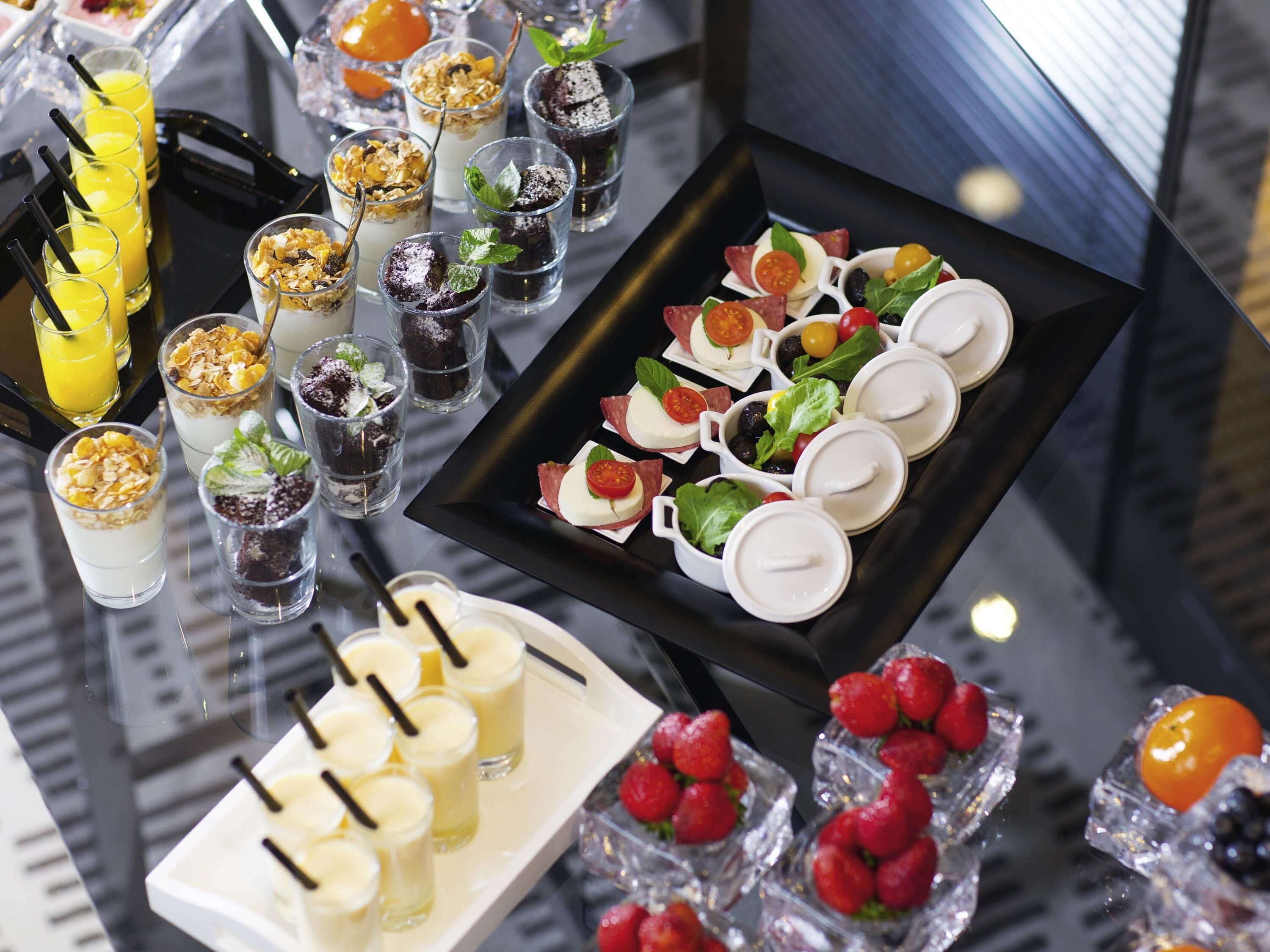 A variety of food and drink options on a glass table, including fruit, cheese, and beverages.