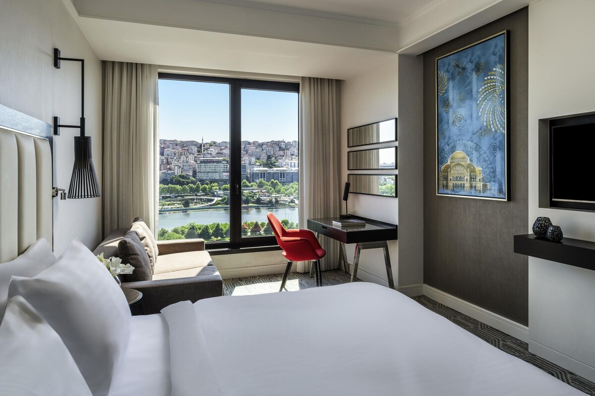 A hotel room with a large bed, a desk with a chair, a TV, a painting, and a view of a cityscape through large windows.