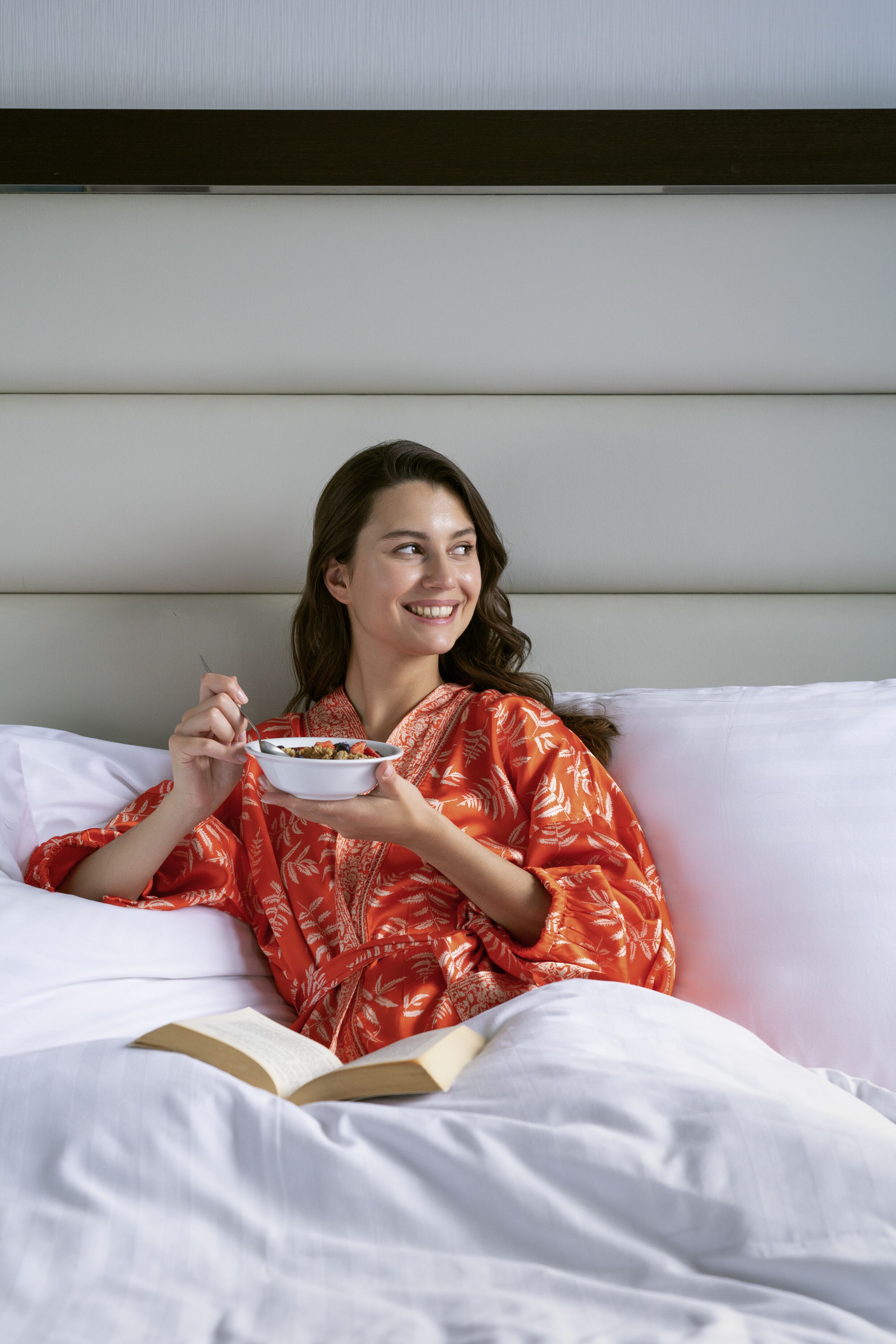 Eine Person sitzt auf einem Bett, neben sich eine Schüssel mit Essen und ein Buch.