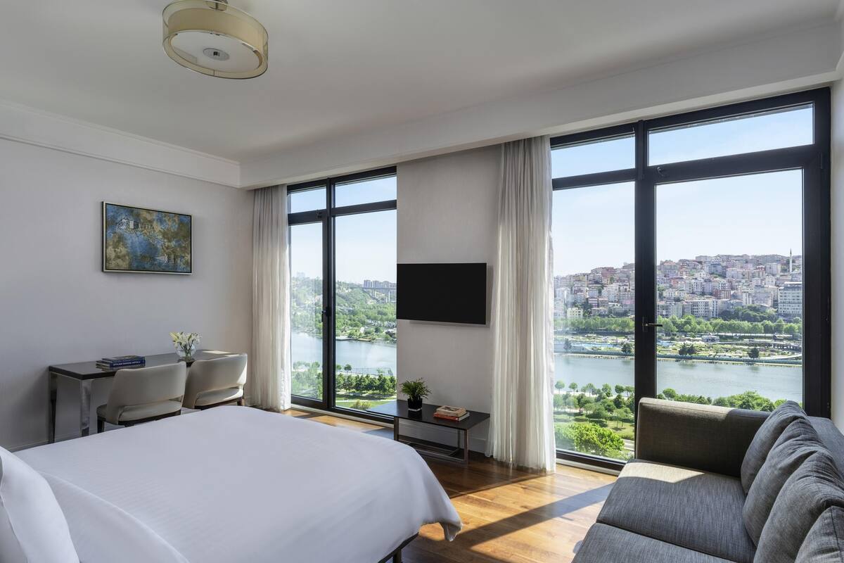 A hotel room with a large bed, a sofa, a desk with a chair, a TV, and a view of a city and river.