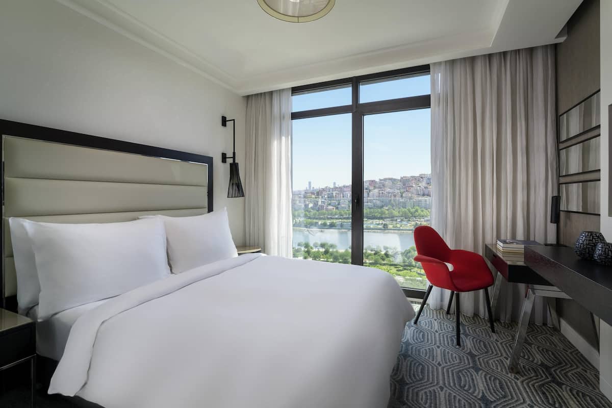A hotel room with a large bed, a desk, a red chair, and a view of a cityscape and river.