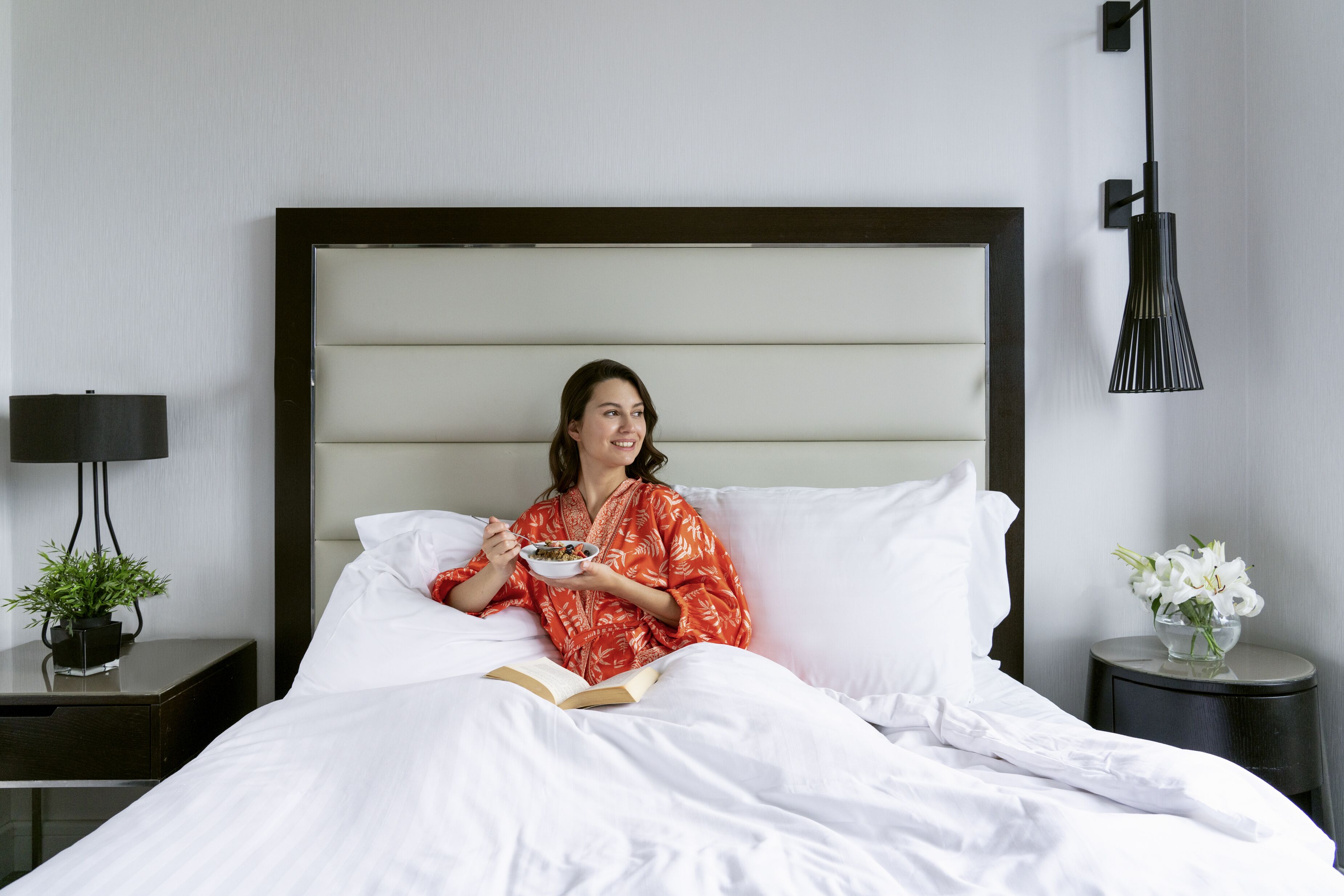 A person sitting on a bed with white bedding, holding a bowl and a book. There is a nightstand with a lamp and a small plant to the left.