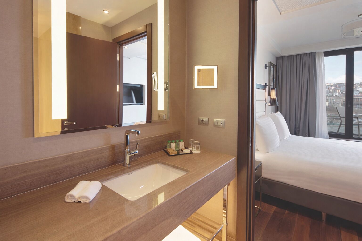 A hotel room with a large mirror, a sink, a neatly made bed, and a view of the city through the window.