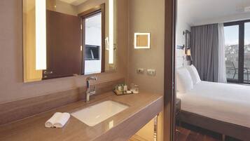 A hotel room with a large mirror, a sink, a neatly made bed, and a view of the city through the window.