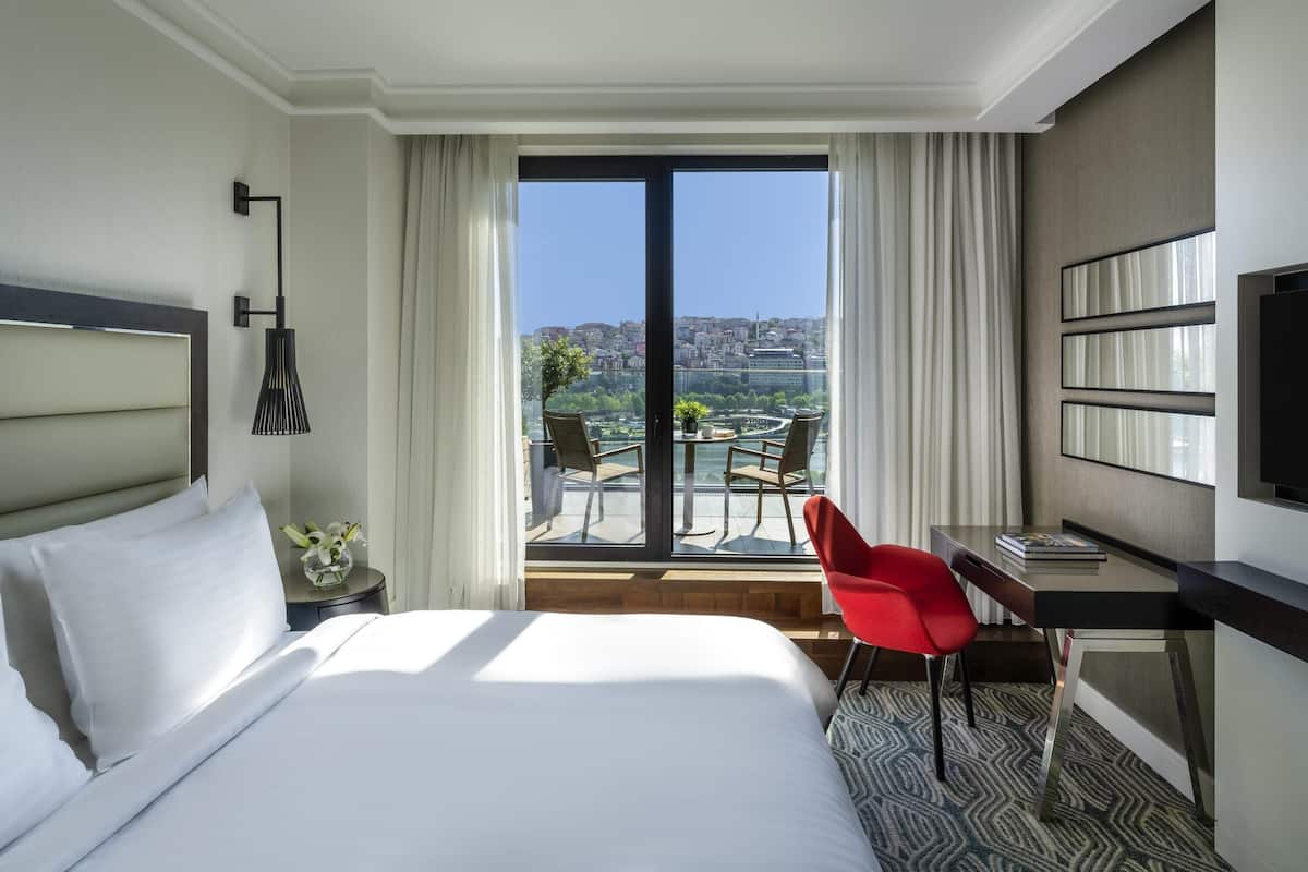A hotel room with a bed, a desk, a chair, a TV, and a balcony with a city view.