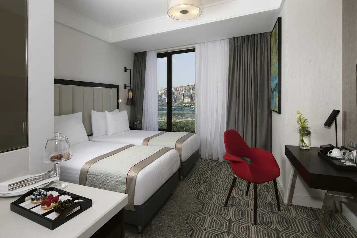 A hotel room with two beds, a desk with a chair, a dining table with a tray of pastries, and a view of a cityscape.