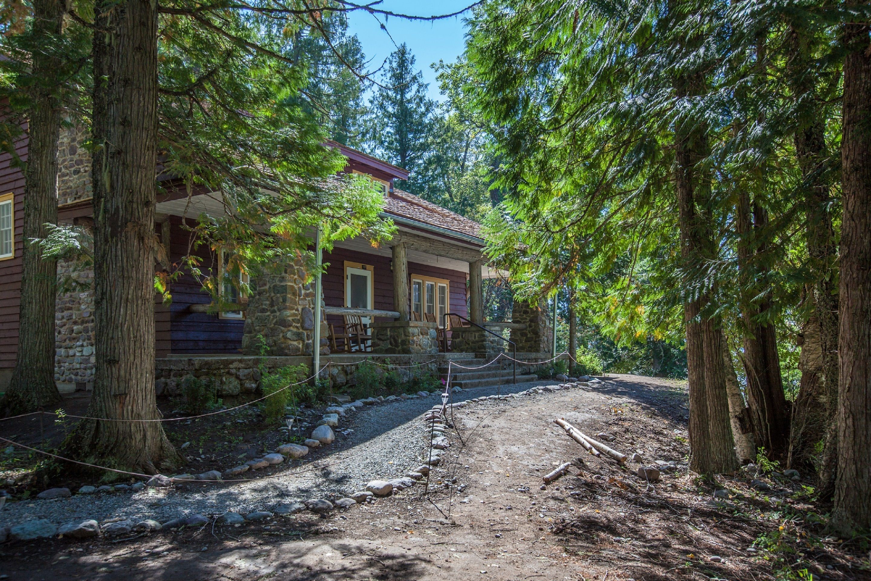Une cabane rustique, dotée de fondations en pierre et d’un revêtement extérieur en bois, entourée de grands arbres et accessible par un chemin de gravier menant à une terrasse en bois.