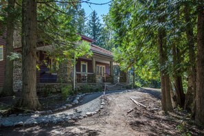 A rustic cabin with a stone foundation and wooden exterior, surrounded by tall trees and a gravel path leading to a wooden deck. - Lake McDonald Lodge - Inside the Park (West Glacier)