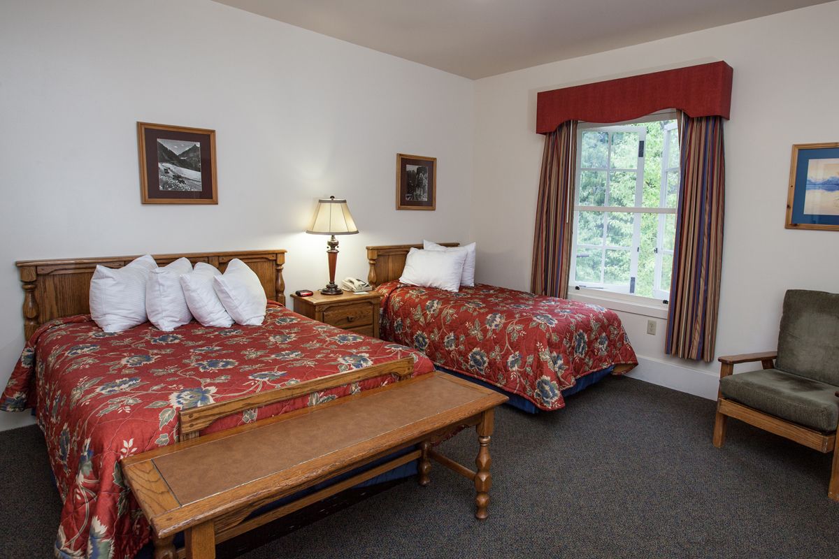 A room with two beds, a bench, a chair, a nightstand with a lamp, and a window with curtains.