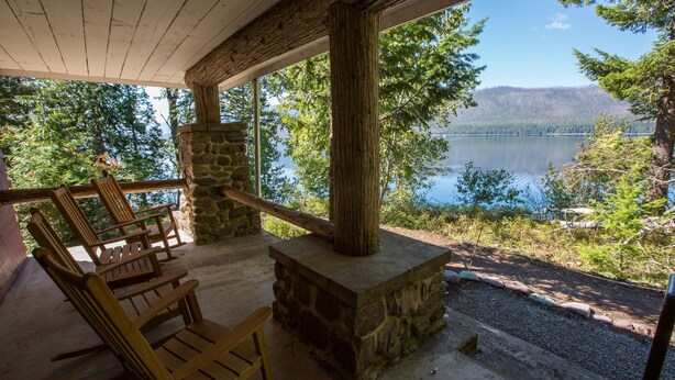 A stone fireplace with a bench and chairs in a covered outdoor area overlooking a lake.