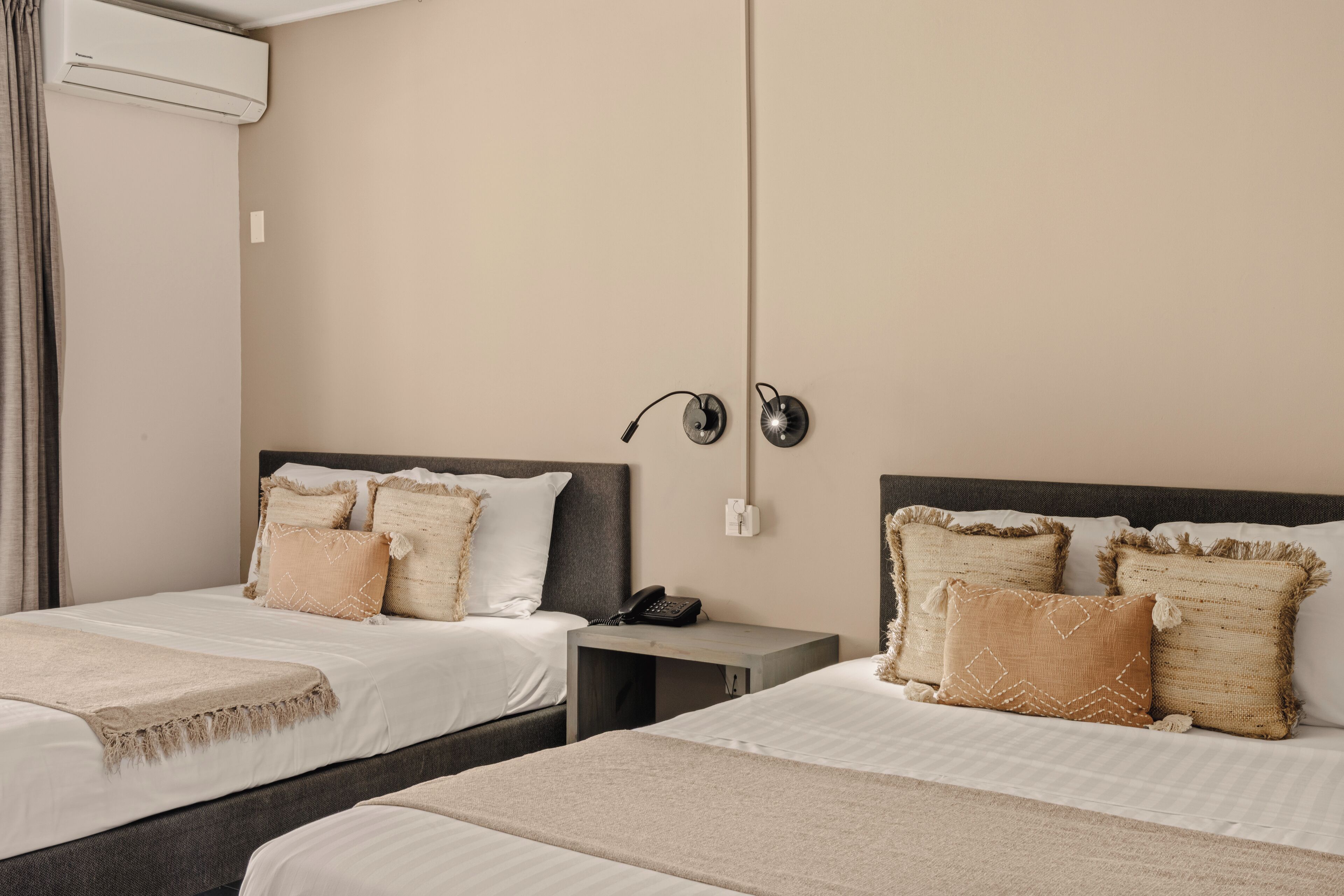 A hotel room with two beds, a bedside table with a phone, and an air conditioning unit on the wall.