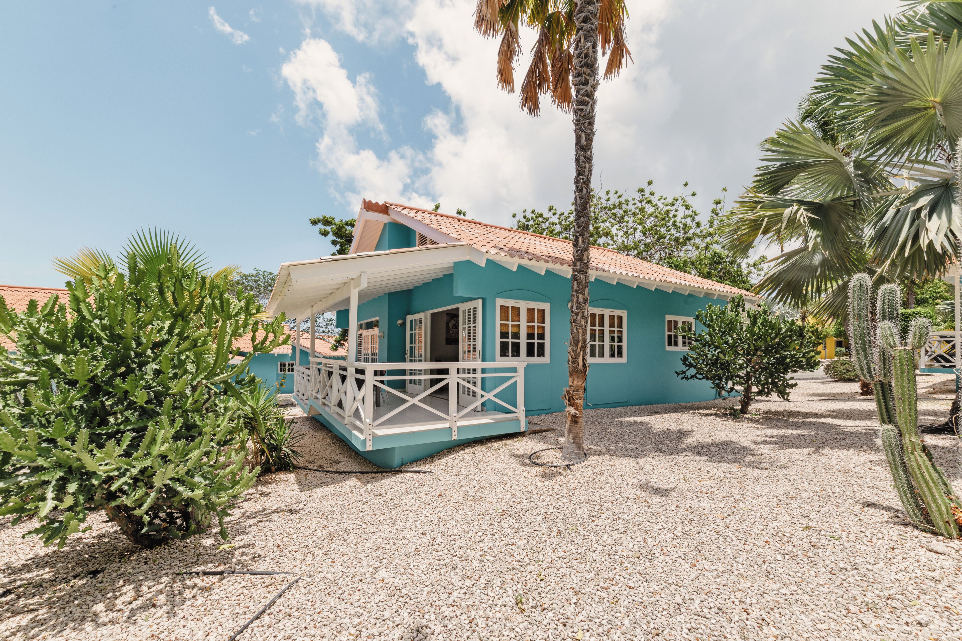 A turquoise building with a white balcony, surrounded by cacti and palm trees.