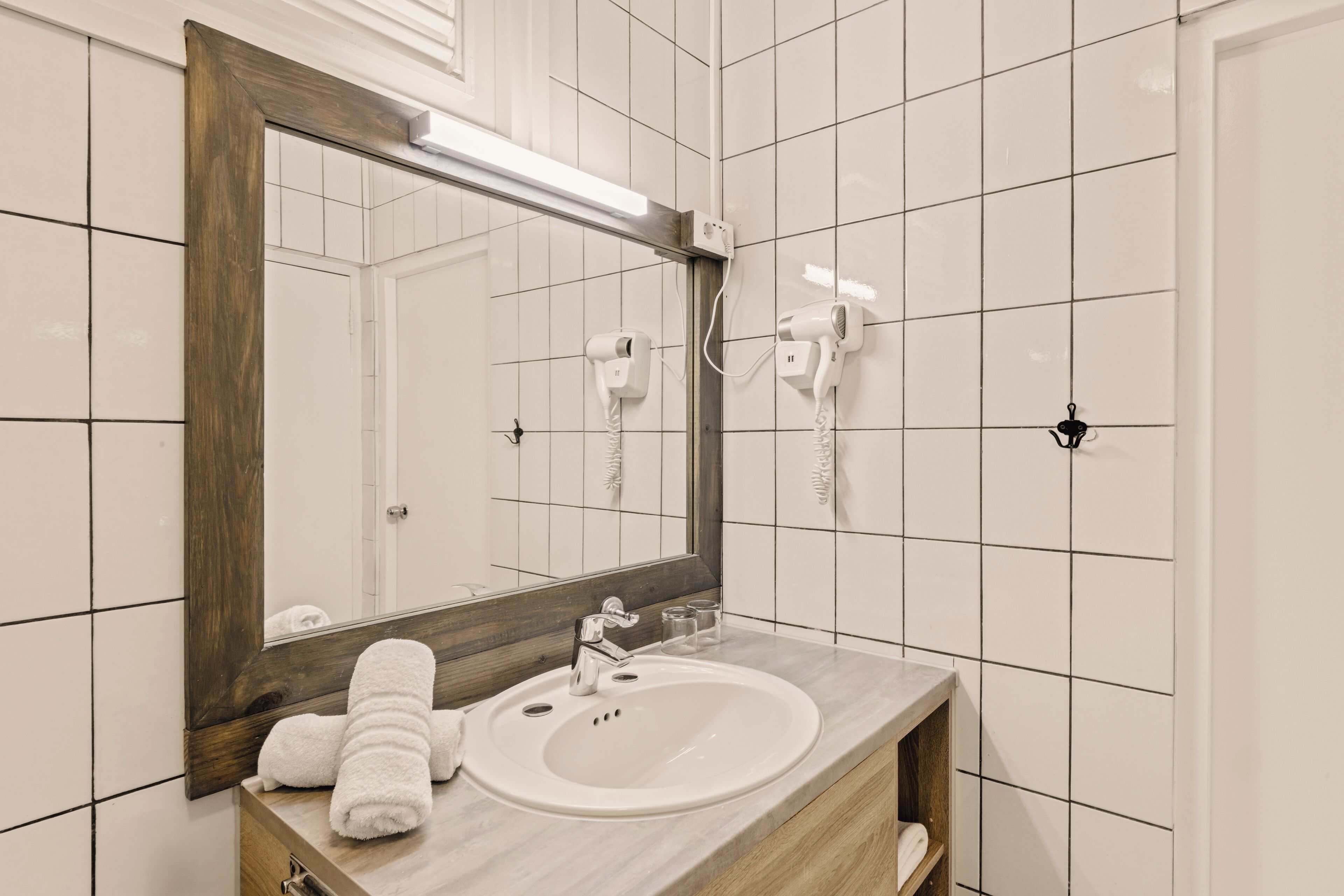 A bathroom with a large mirror, a sink, two hair dryers mounted on the wall, and a towel on the counter.