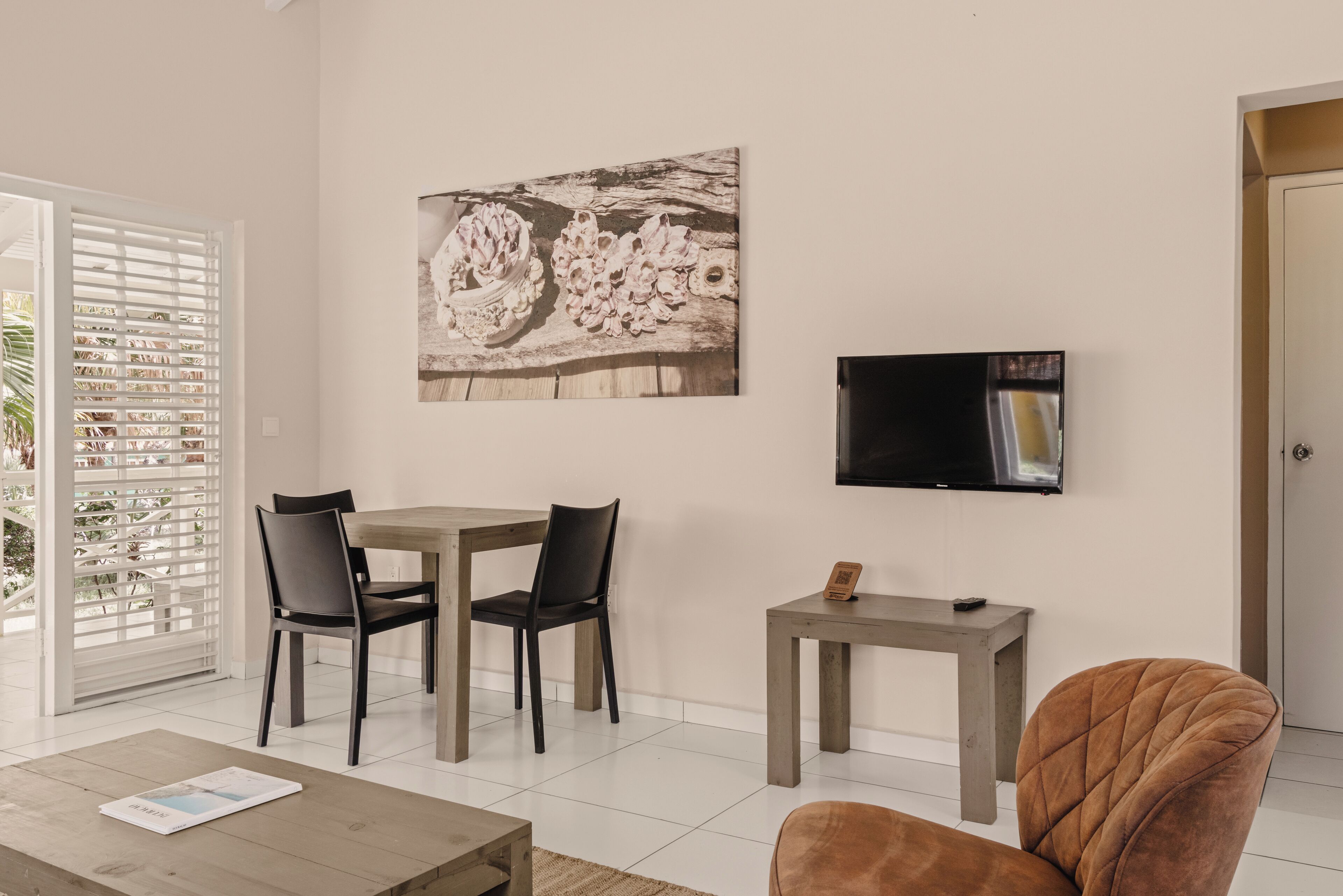 A living room with a dining table, two chairs, a coffee table, a TV mounted on the wall, and a door to the right.