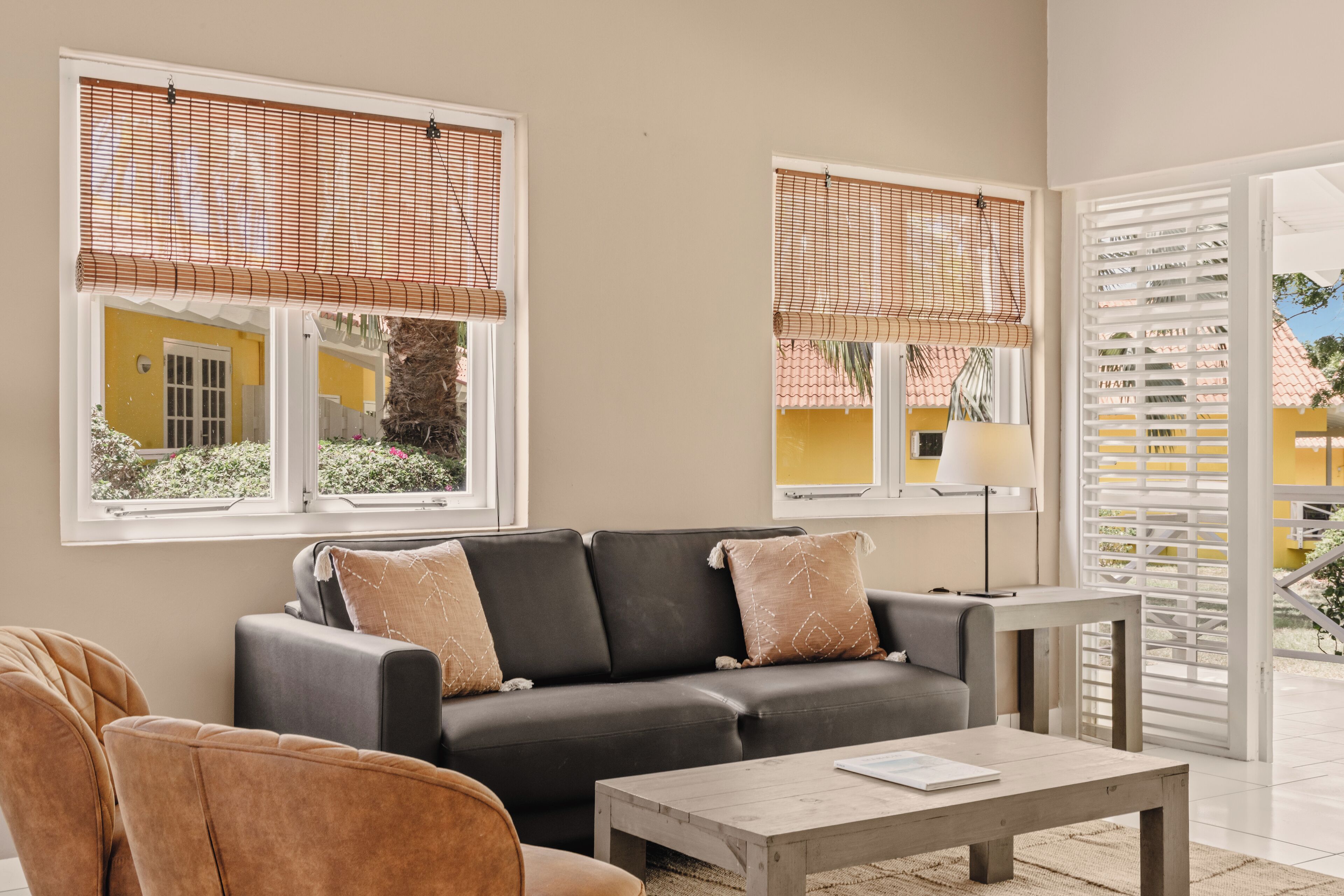 Una sala de estar con un sofá, sillones, una mesa de centro y una lámpara. Hay ventanas con persianas y vistas al exterior.